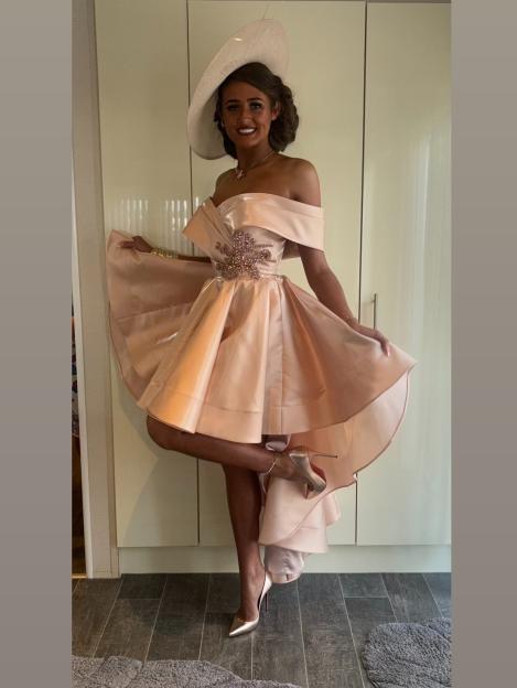 A woman in a peach-colored off-the-shoulder dress with a wide-brimmed hat poses with one leg bent.
