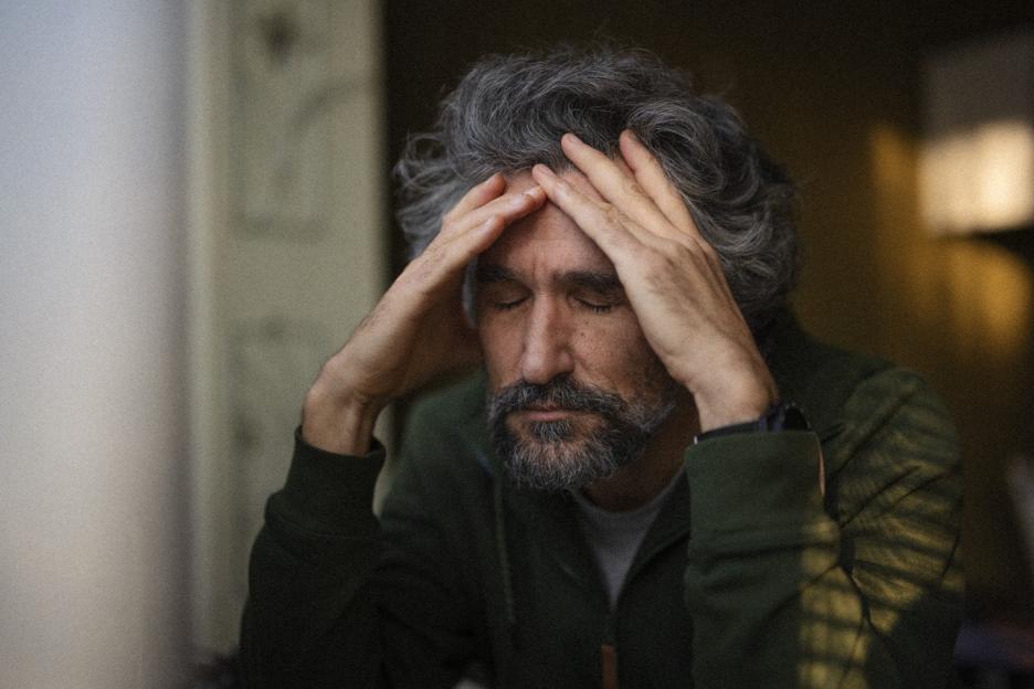 Mature man with gray hair and beard, wearing a green jacket, holding his head in his hands with closed eyes, appearing stressed or meditating.