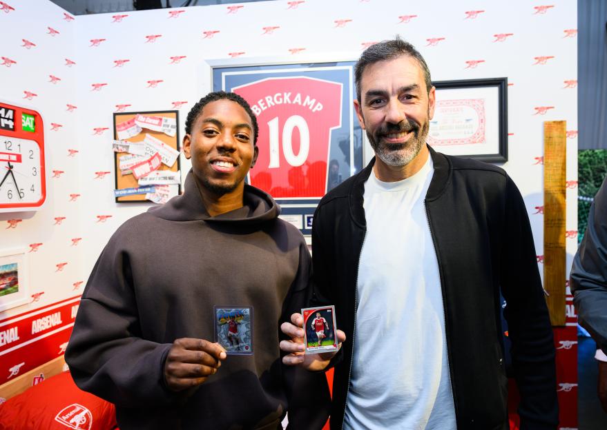 Two men, including former footballer Robert Pires, holding Topps Chrome trading cards of Myles Lewis-Skelly in a room decorated with Arsenal Football Club memorabilia.