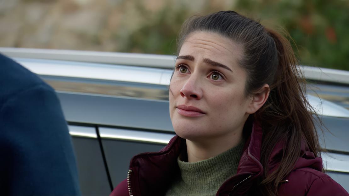 A woman with dark hair tied back, wearing a green turtleneck and a maroon jacket, looks up with a distressed expression.