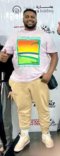 Full-body shot of smiling Abdullah Hassan wearing a pink t-shirt with "Calabasas" and a landscape graphic, tan sweatpants, and light-colored sneakers.