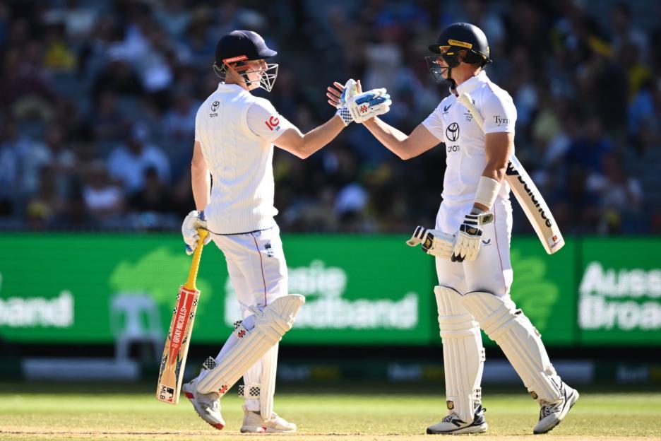 Two cricketers from England, dressed in white, shake hands in the middle of a game, with a crowd in the background.