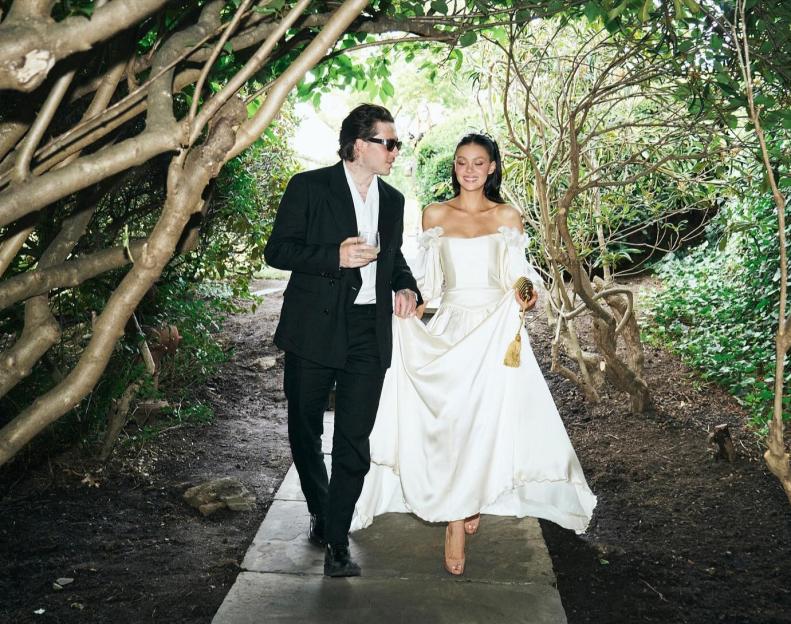 A man in a black suit holding a drink and a woman in a white off-the-shoulder dress walking together on a path under intertwined branches.