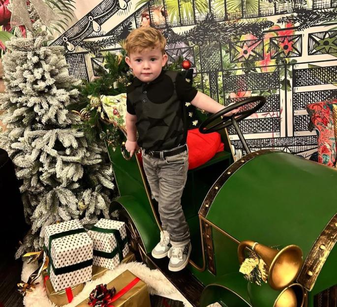 Peter Maughan, also known as Peter Rabbit, standing in a green toy car next to a Christmas tree and wrapped presents.