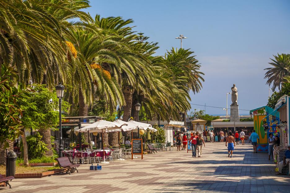 A wide pedestrian promenade in Tarifa, Spain, with palm trees, outdoor cafes, and people walking, under a clear blue sky.
