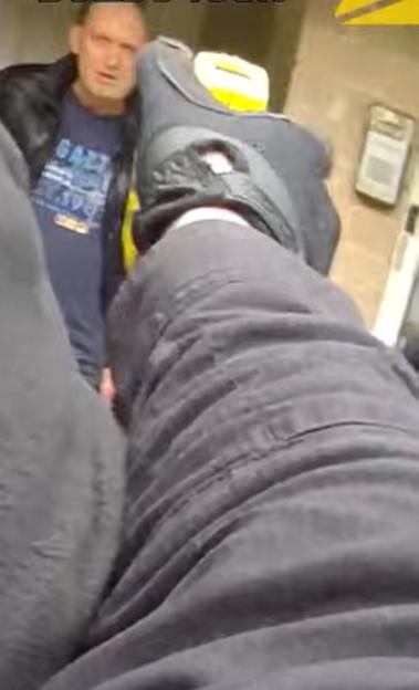 An individual in dark clothing and a black shoe seen from a first-person perspective, with a man wearing a dark t-shirt and jacket in the background.