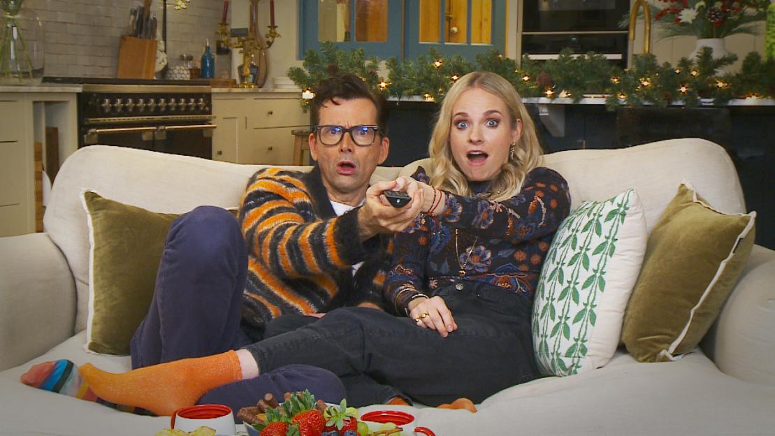 A man and a woman on a couch looking surprised and pointing a remote control forward.
