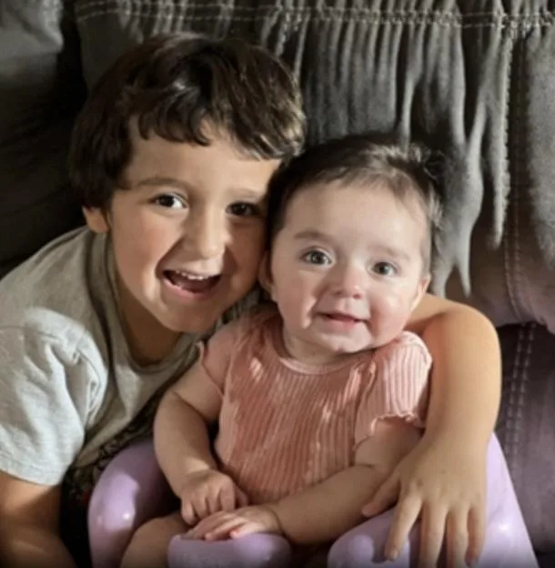 A boy smiles and hugs a baby in a lavender chair.