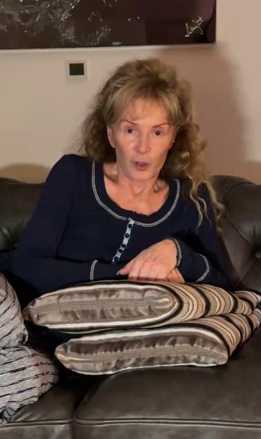 A woman with shoulder-length curly hair sits on a brown leather couch with her hands clasped, speaking.