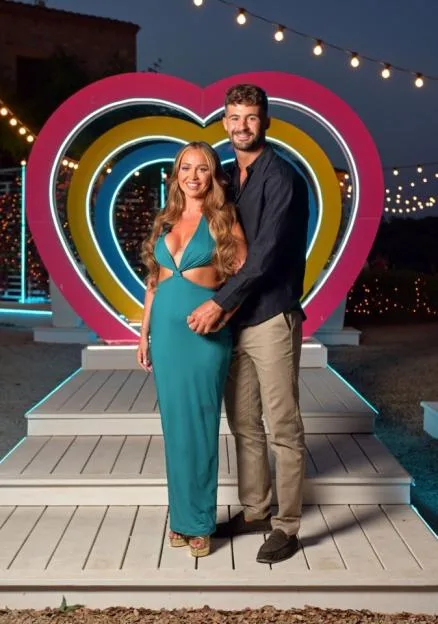 A couple stands together on a lit stage in front of a giant heart-shaped display on Love Island.