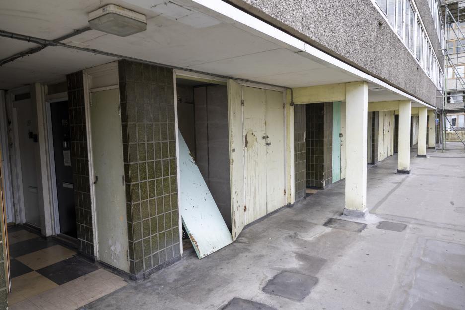 Exterior of the Aylesbury Estate in Walworth, South London, showing multiple doors to flats, one door ajar with a board leaning inside, and scaffolding visible to the right.