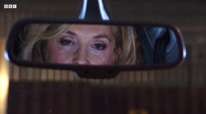 A woman with blonde hair and blue eyes reflected in a car's rearview mirror.