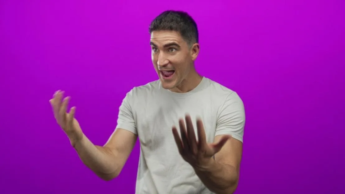 A man in a white t-shirt communicates with enthusiastic hand gestures in front of a purple background.