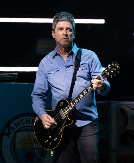 Noel Gallagher performing on stage, wearing a denim shirt and playing a black Gibson electric guitar.