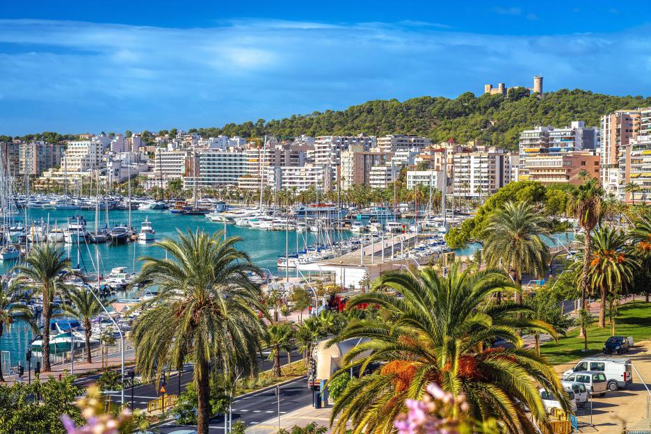 Palma de Mallorca yachting harbor waterfront with boats, city buildings, and a distant castle on a hill.