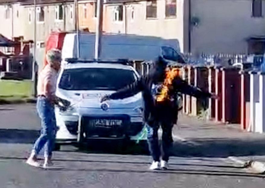 A man in a dark hoodie with a bright orange flame on his chest pulls a white police car with a rope, while another man in a light shirt and jeans walks beside him.