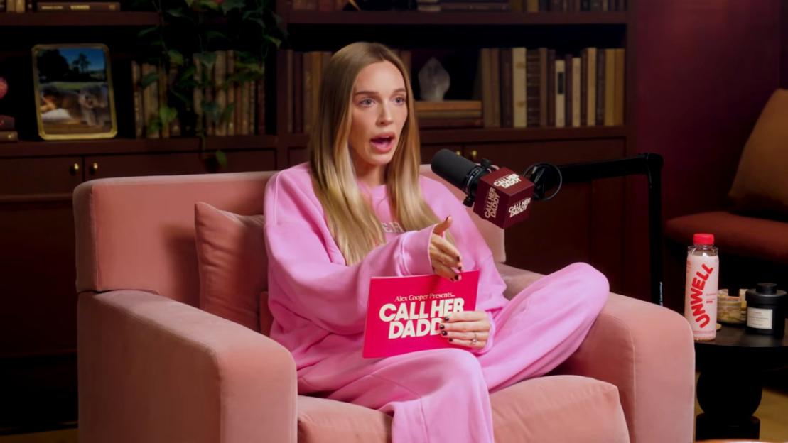 A woman wearing a pink tracksuit is seated in a pink armchair, holding a red card that reads "Call Her Daddy" with a microphone next to her, indicating a podcast recording.