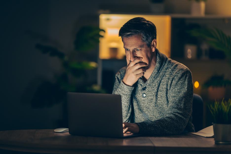A man sits at a desk in a dimly lit room, intensely focused on his laptop, with his hand resting on his chin.