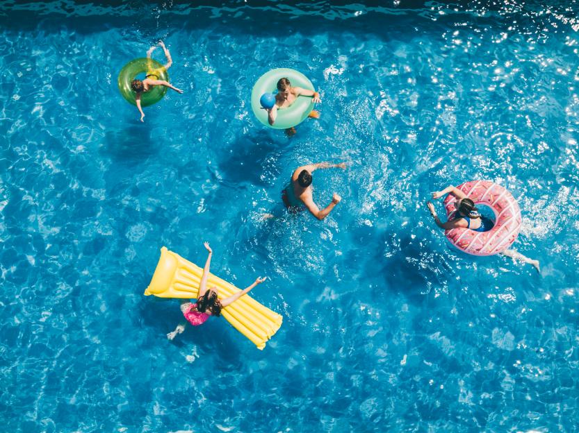 Five people swimming and relaxing in a blue pool with floats.