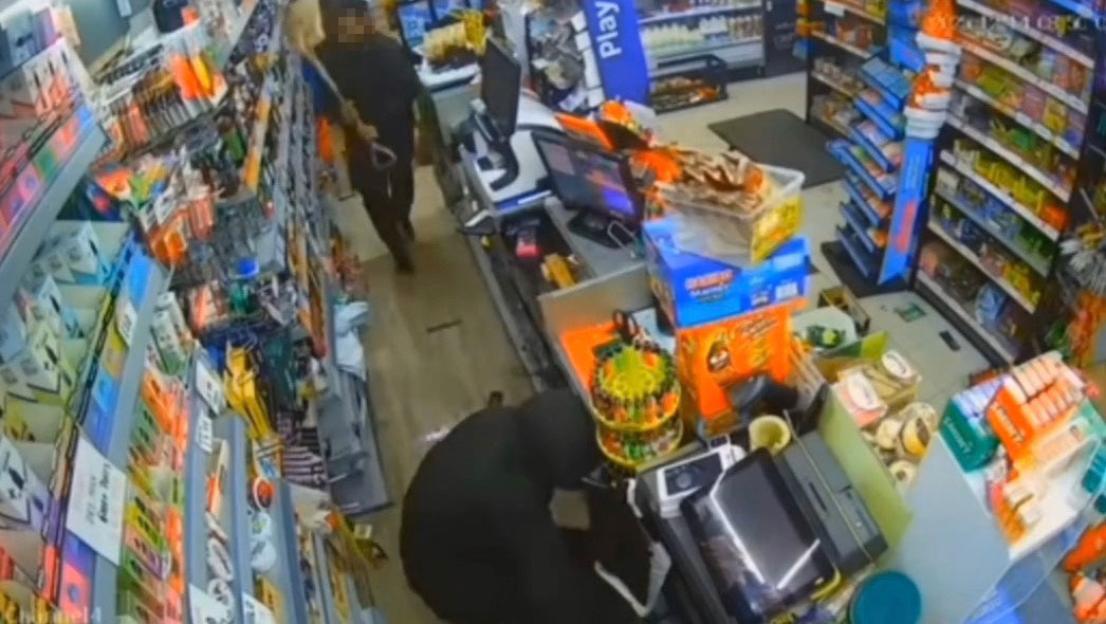 A shopkeeper with a shovel and a hooded individual confronting each other in a store aisle.