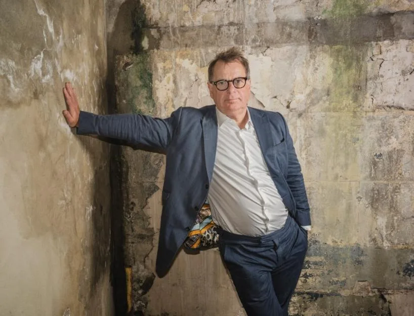 A man in a blue suit and glasses leans against a weathered, peeling concrete wall.