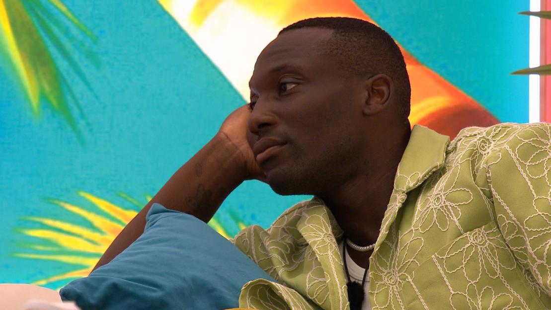 Ope from Love Island looking on with his hand resting on a blue pillow.