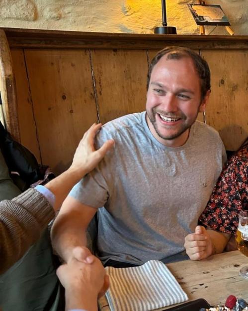 A man in a gray t-shirt shakes hands with another person at a wooden table in a pub.