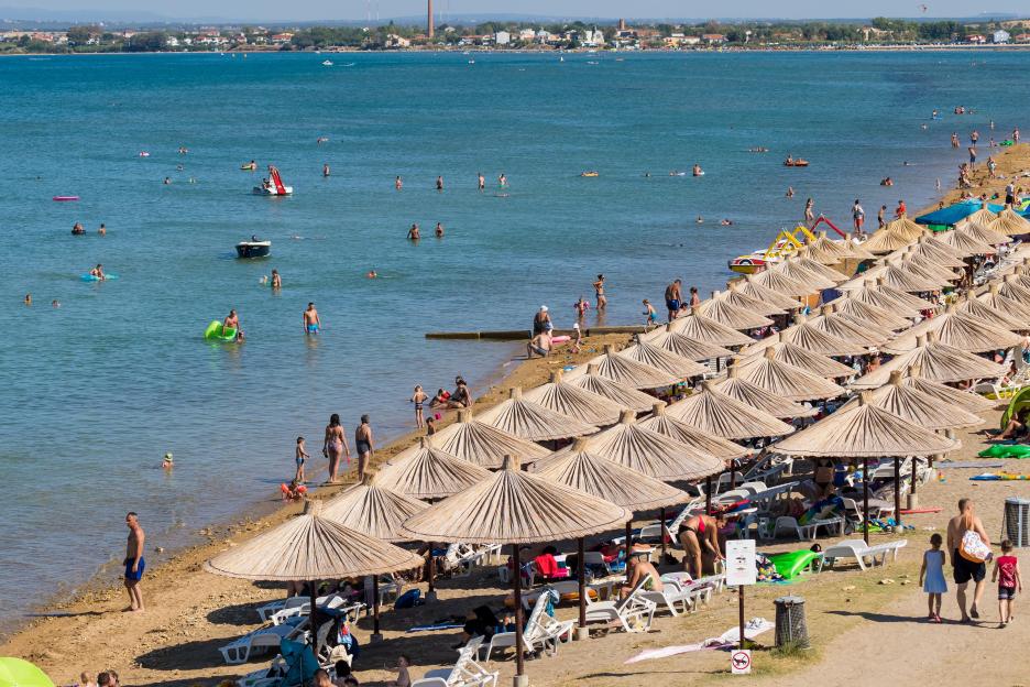 Beach view at Nin, Dalmatia, Croatia, showing people swimming in the sea and relaxing under straw umbrellas on the sand.