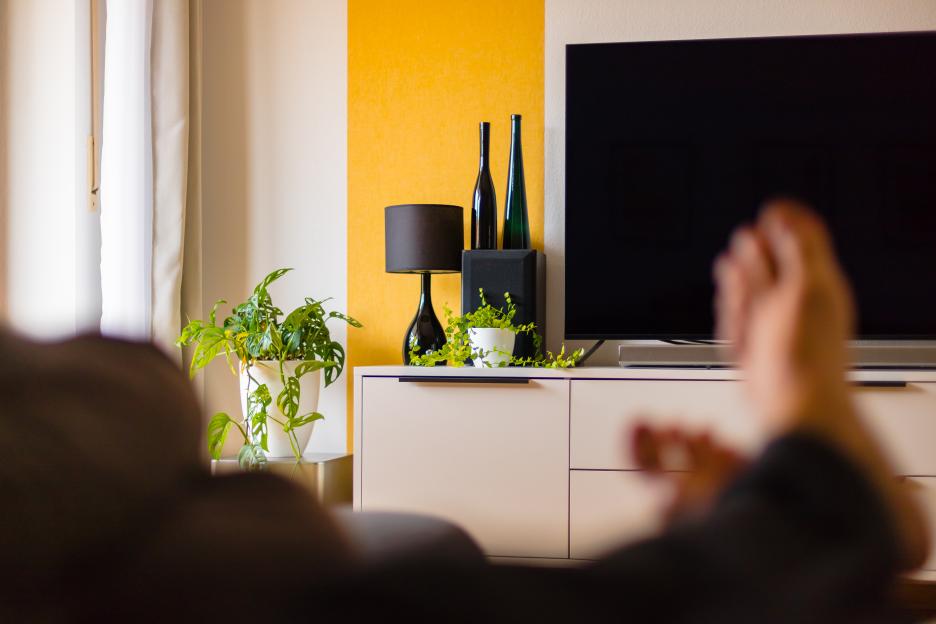 A person sitting on a sofa in a living room with their feet crossed and propped up, with a TV, plants, and yellow wallpaper visible in the background.