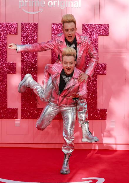 Jedward in pink and silver outfits posing on a pink and glittery background.