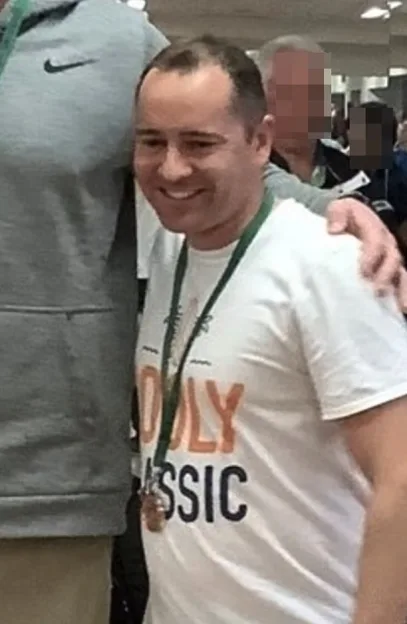 Christopher Gerald Savage smiling while wearing a white t-shirt and a lanyard with a medal.