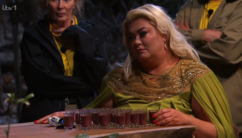 A woman in a green and gold dress looks at a tray of small glasses filled with a dark liquid.