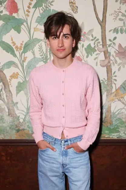Sebastian Croft in a pink cardigan and jeans, posing against a floral wallpaper.