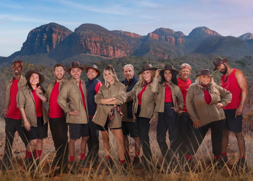 Cast of "I'm a Celebrity... South Africa" smiling, dressed in khaki and red, against a mountainous backdrop.