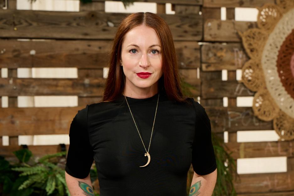 A woman with long red hair, wearing a black dress and a crescent-shaped pendant, stands with her hands on her hips.