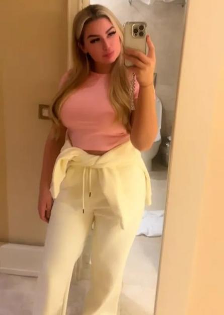 A woman with long blonde hair and a pink top takes a mirror selfie.