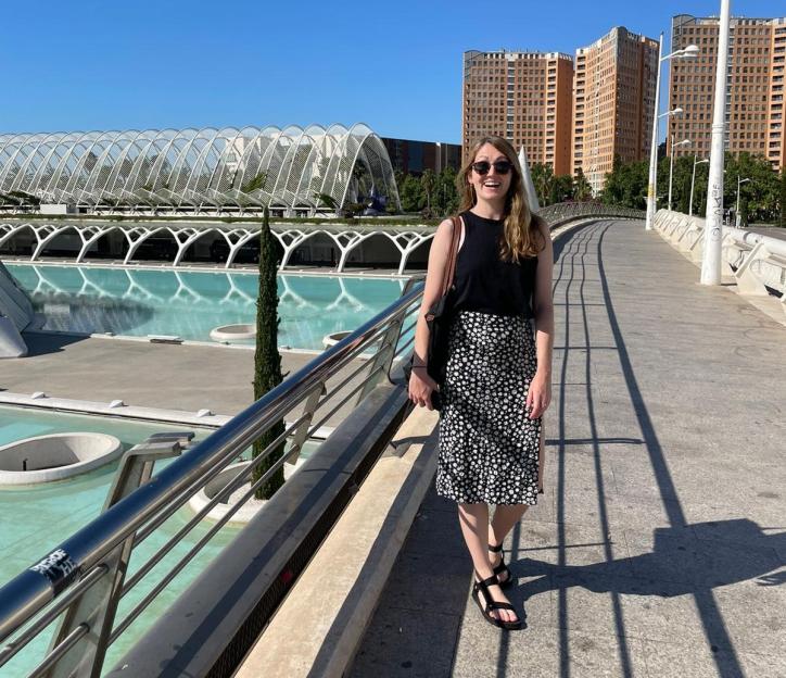 A young woman smiling on a bridge with modern architecture and buildings in the background.