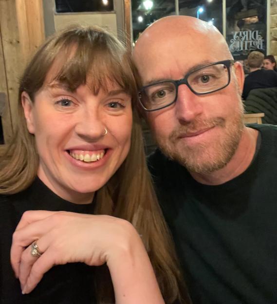 A smiling woman with long brown hair and a man with glasses and a beard in a cozy, dimly lit interior.