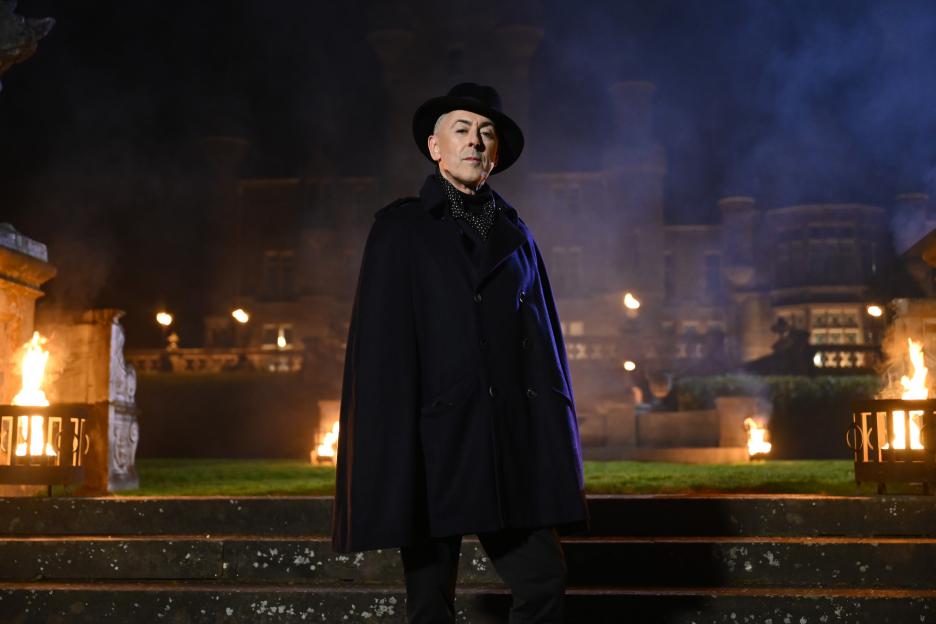 Alan Cumming in a dark blue cape and hat, standing in front of a castle with fire pits.