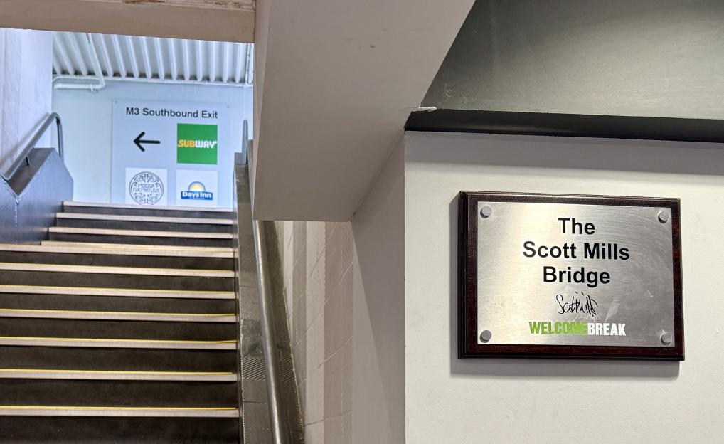 A plaque on a wall for "The Scott Mills Bridge" at Welcome Break Fleet Services, with a stairway and signs for M3 Southbound Exit, Subway, Pizza Express, and Days Inn in the background.