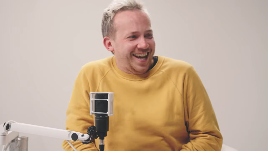 A man with blonde hair and a yellow sweatshirt smiling, next to a microphone.
