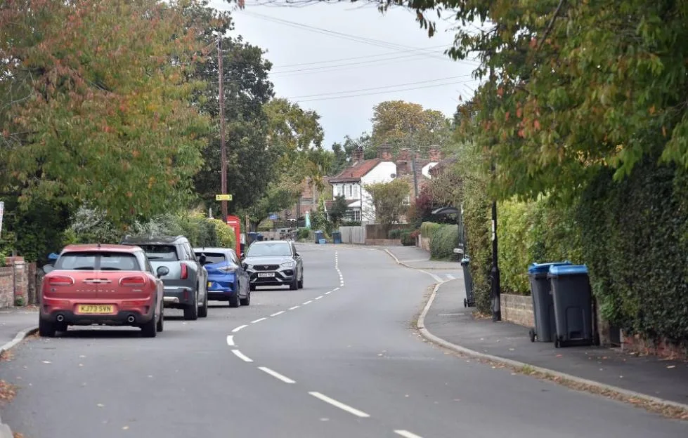 A quiet street in the village of Linton-on-Ouse, flanked by parked cars, houses, and trees.