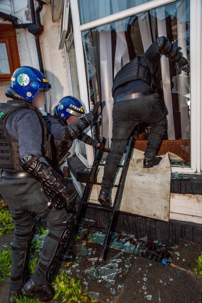 Police officers storming a house through a broken window using a small ladder during a drug raid.