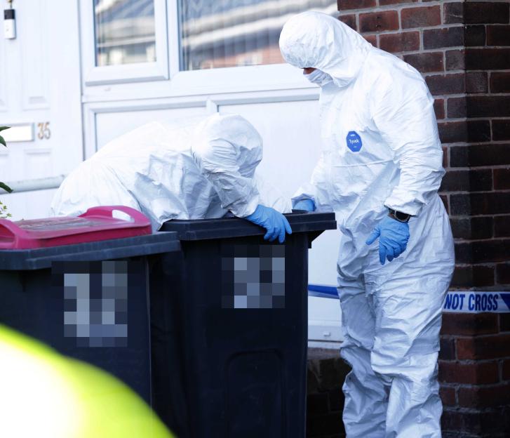 Two forensic officers in white suits and blue gloves investigate two black bins at a crime scene.
