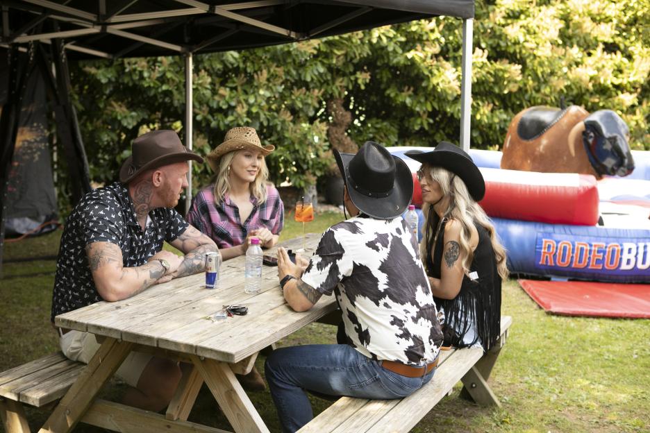 Four people wearing cowboy hats sit around a picnic table, with a mechanical bull ride in the background.
