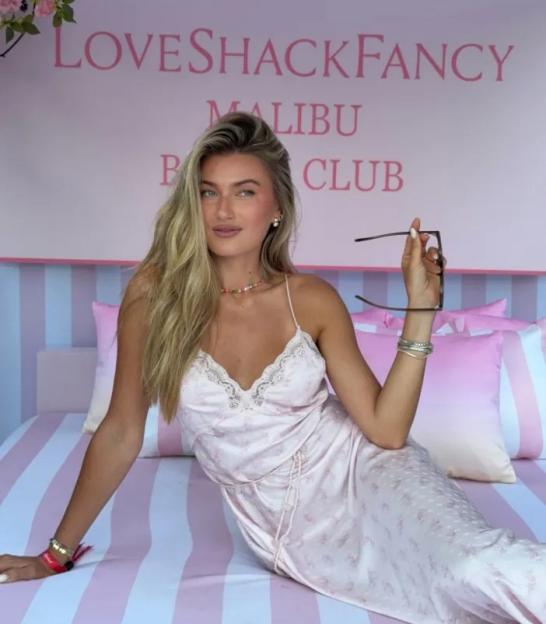 Woman in a pink slip dress holding sunglasses on a bed with a "LoveShackFancy Malibu Beach Club" sign in the background.
