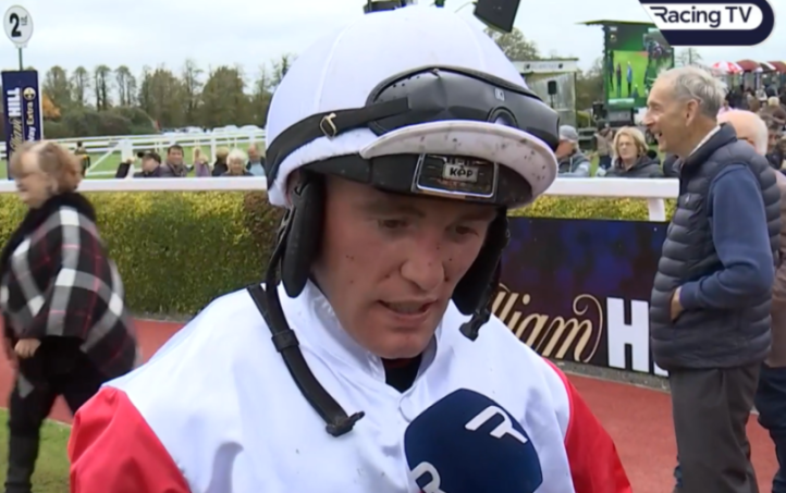A jockey in a white helmet and red and white shirt is being interviewed, holding a blue microphone.