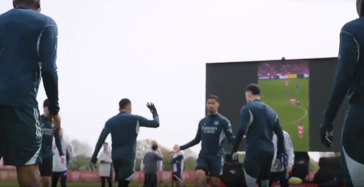 Football players in blue and black uniforms watch a game on a giant screen.