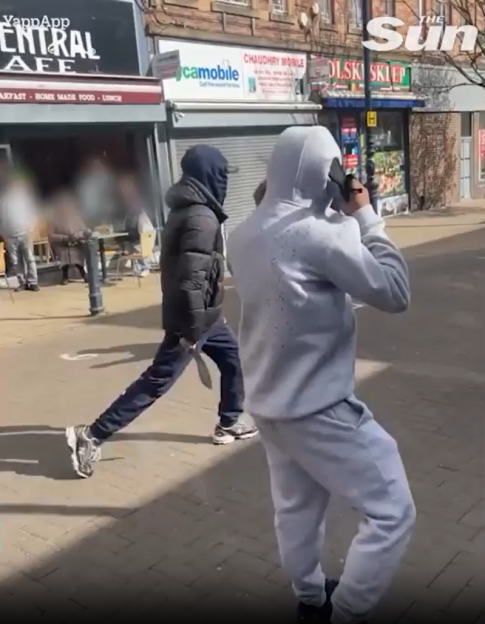 A person in a balaclava and dark jacket holding a knife, walking past a person in a grey tracksuit filming them.