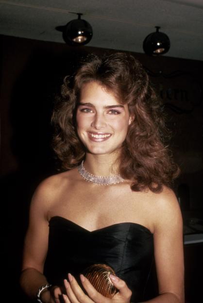Brooke Shields smiling in a black strapless dress and a rhinestone choker, holding a small gold clutch.